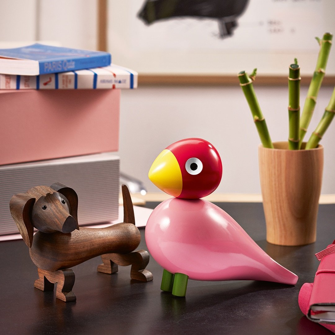Colorful wooden bird figurine in pink and yellow, paired with a brown dog sculpture and green bamboo in a minimalist setting.