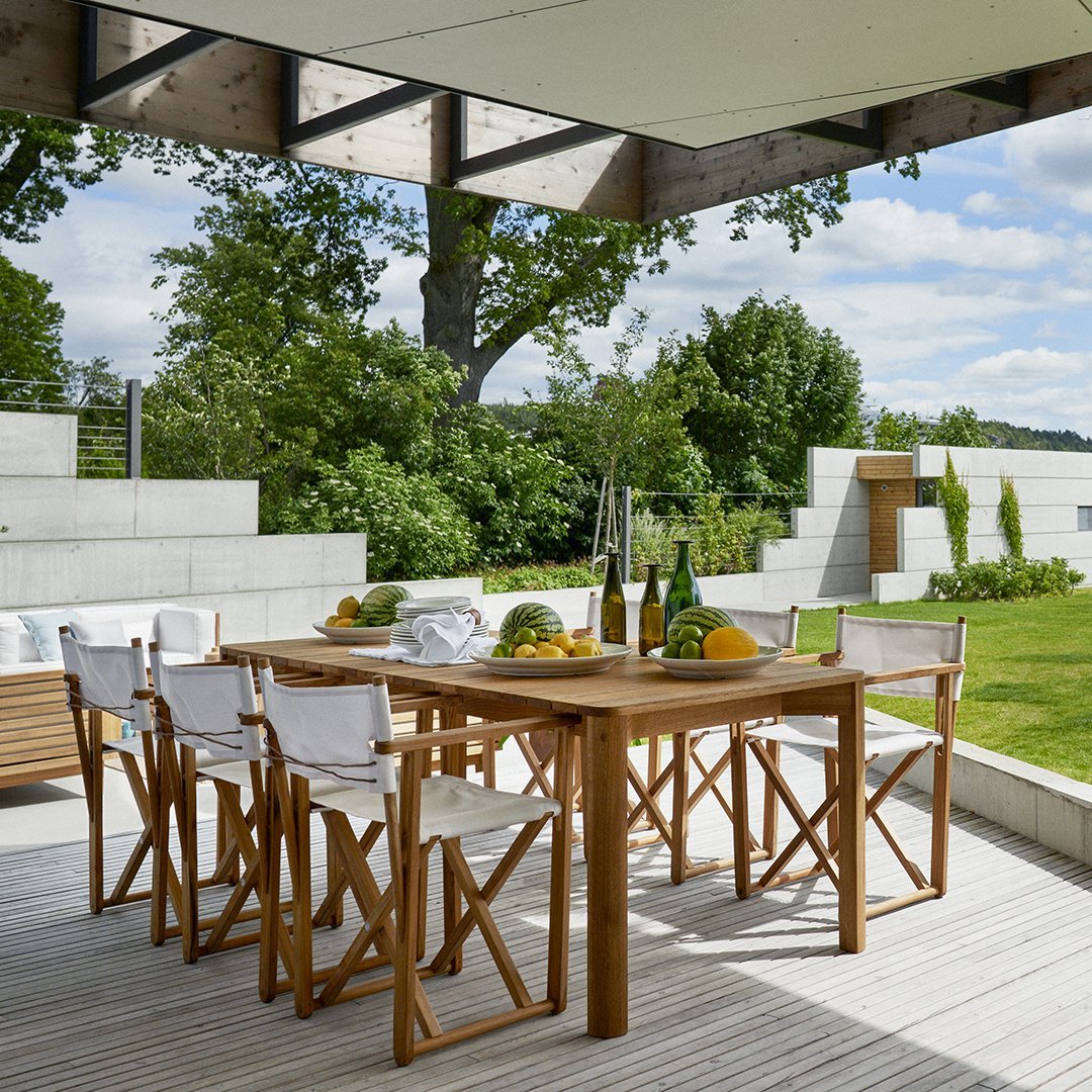 "Natural wood dining table with white fabric chairs, set against a green garden backdrop and blue sky."