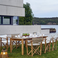 "Elegant wooden dining table with light beige and white accents, set against a serene outdoor backdrop of green and gray."