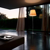Modern floor lamp with a soft beige shade, sleek silver base, and warm ambient lighting, surrounded by sheer curtains.
