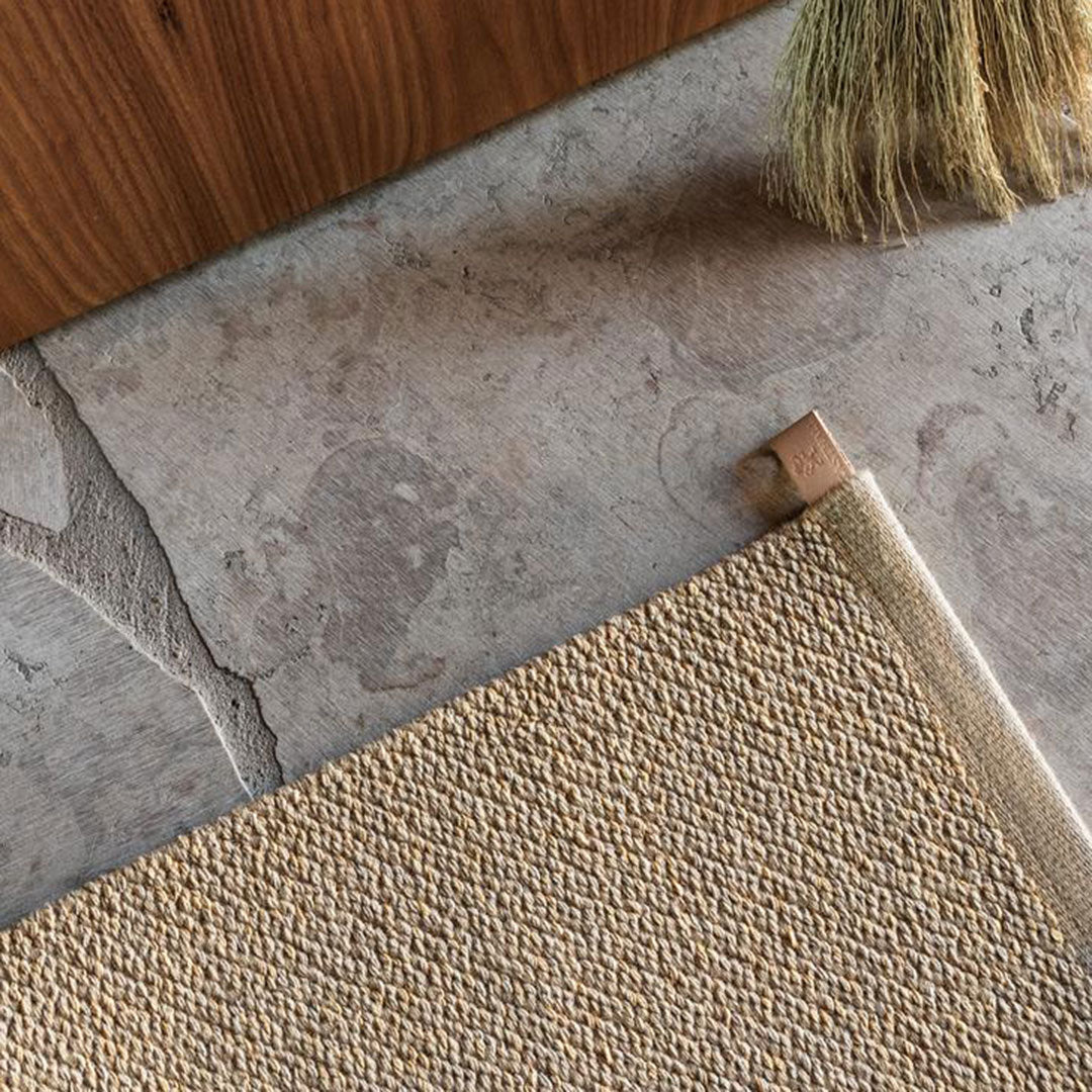 Textured beige rug corner on smooth gray stone floor, accented by light wood and natural brush.