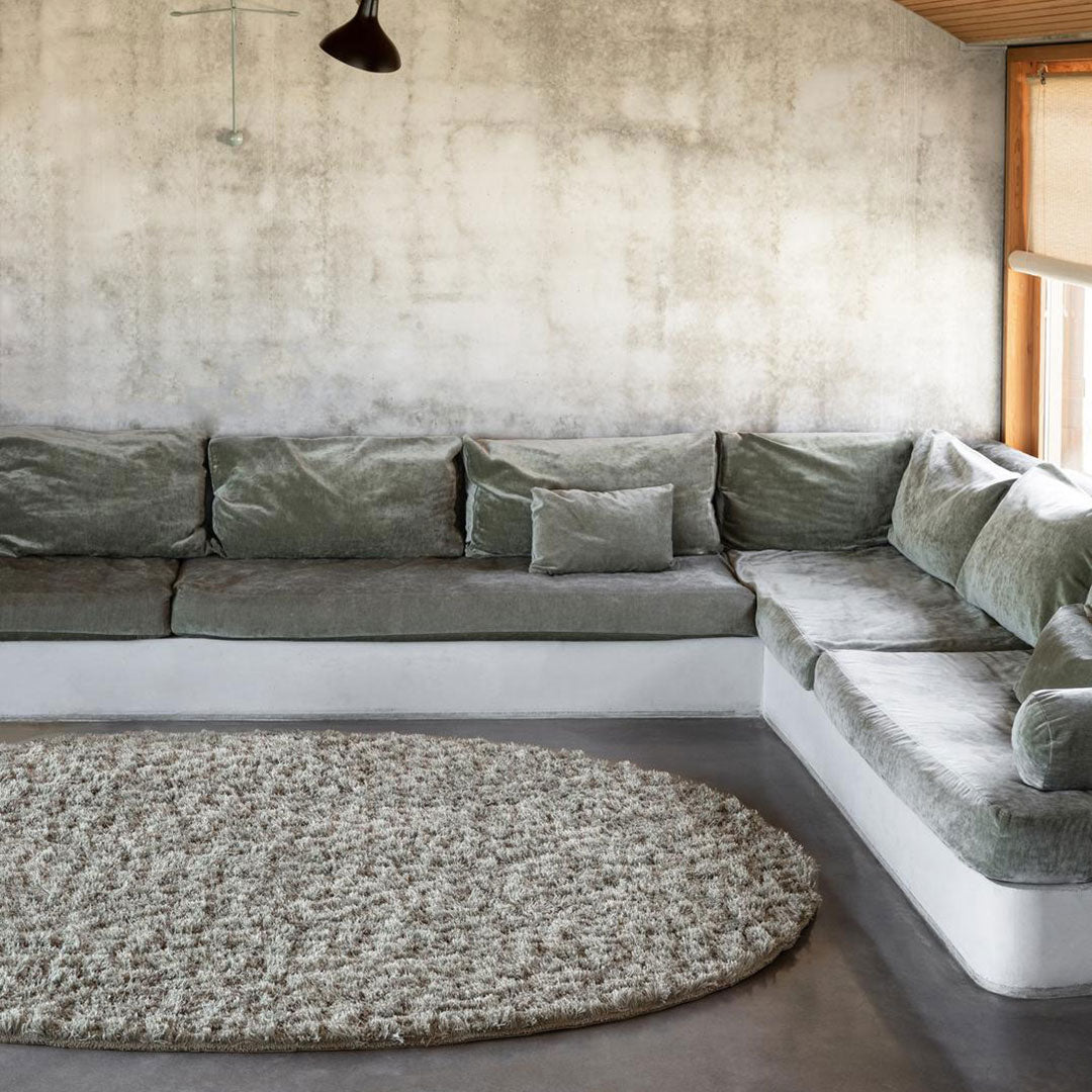 Green velvet sectional sofa with gray cushions and a beige patterned round rug on a concrete floor.