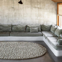 Green velvet sectional sofa with gray cushions and a beige patterned round rug on a concrete floor.