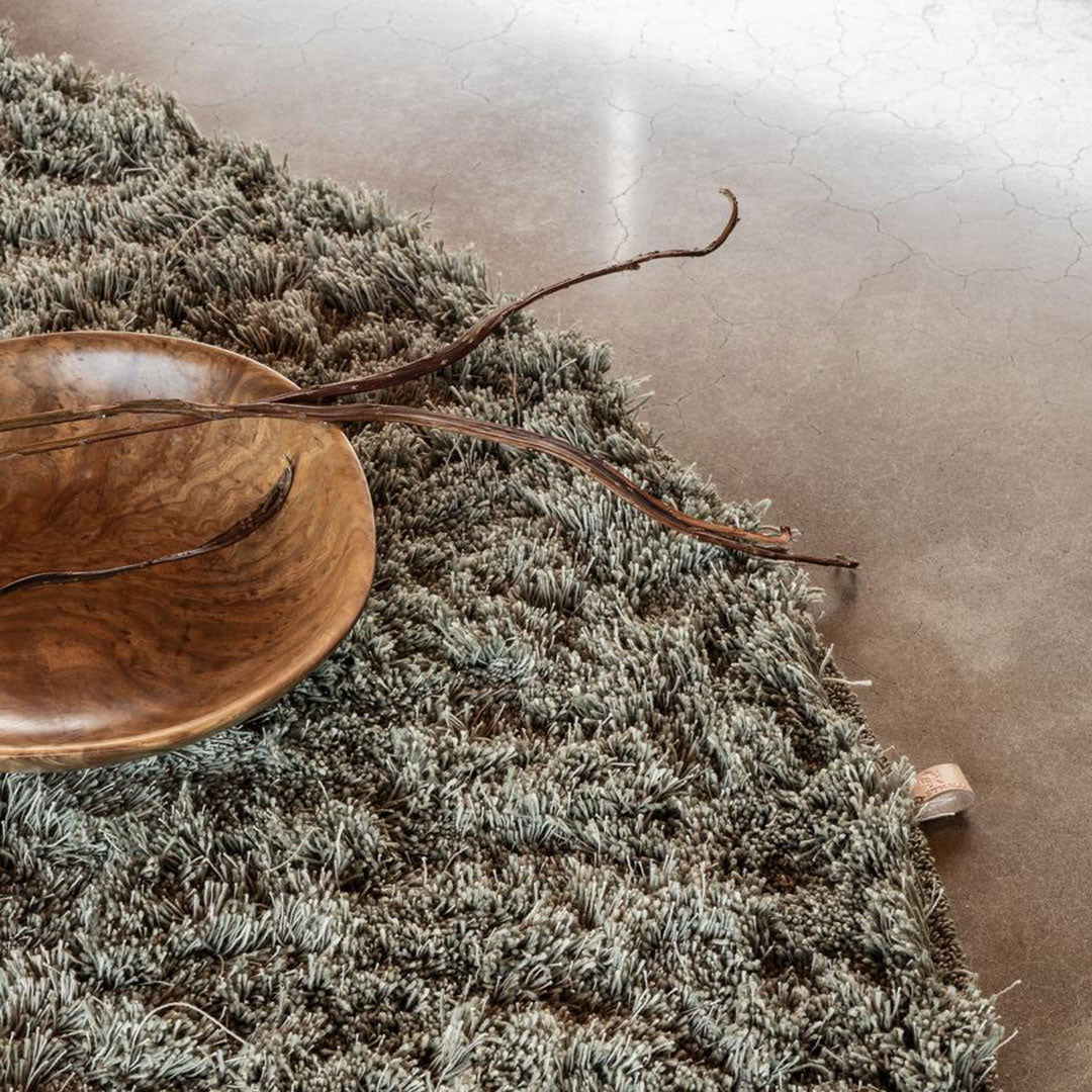 Textured green rug with soft, muted tones, featuring a natural, organic pattern. Stylish round wooden bowl accent.
