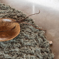 Textured green rug with soft, muted tones, featuring a natural, organic pattern. Stylish round wooden bowl accent.