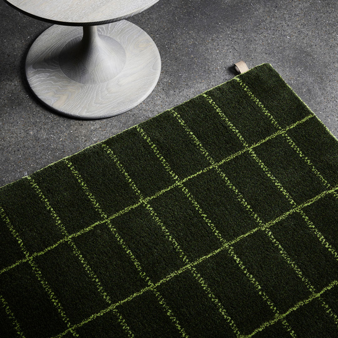 Green plaid rug with contrasting light green grid pattern on dark green background, showcased on a gray surface.