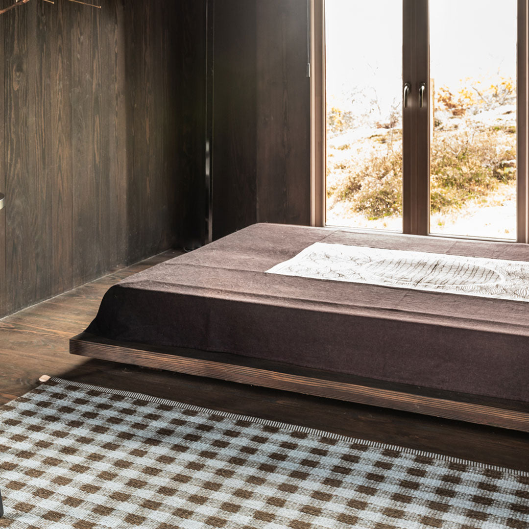 Brown bedspread with a textured finish layered over a beige and brown checkered rug. Natural light enhances earthy tones.