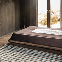 Brown bedspread with a textured finish layered over a beige and brown checkered rug. Natural light enhances earthy tones.