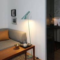Modern floor lamp in teal, paired with a gray sofa and brown accent pillow, set against a minimalist white wall.