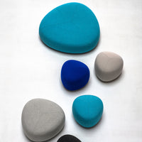 Colorful, smooth, pebble-shaped cushions in shades of teal, blue, gray, and black, featuring modern, minimalist design.