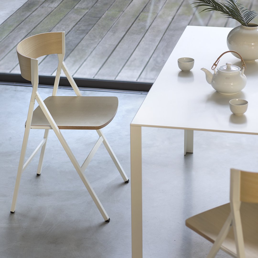 Modern white folding chair with light wood accents, set against a sleek gray floor and minimalist table decor.
