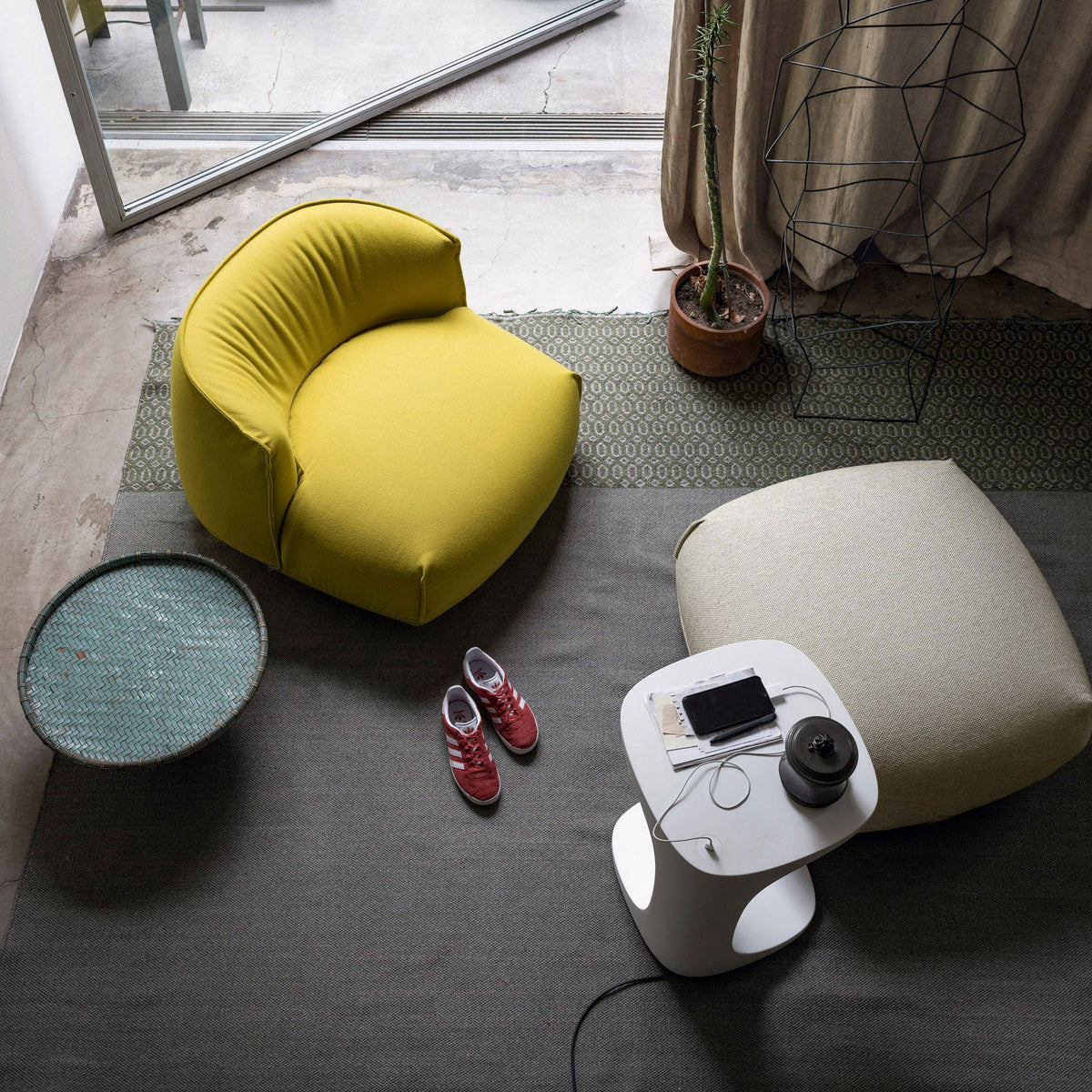 Bright yellow curvy chair on a green patterned rug beside a neutral-colored ottoman and a blue textured table.