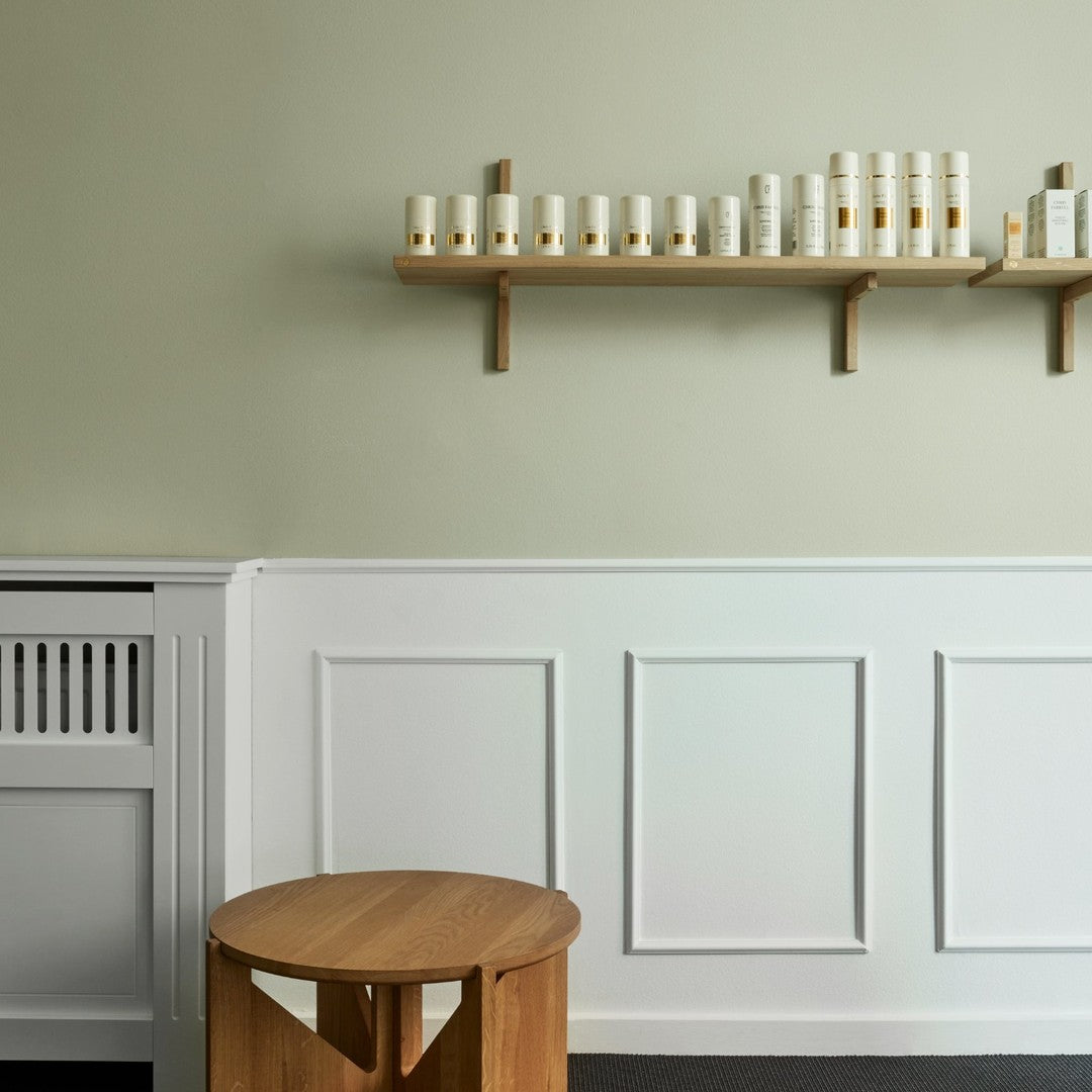Wooden stool in front of a muted green wall, with a white paneled section and a natural wood shelf displaying candles.