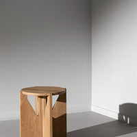 Light wood round stool with geometric cutout, contrasted against a minimalist gray wall and floor.