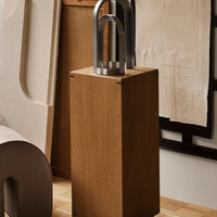 Natural wood grain pedestal with a sleek metallic sculpture on top, set against a neutral background.