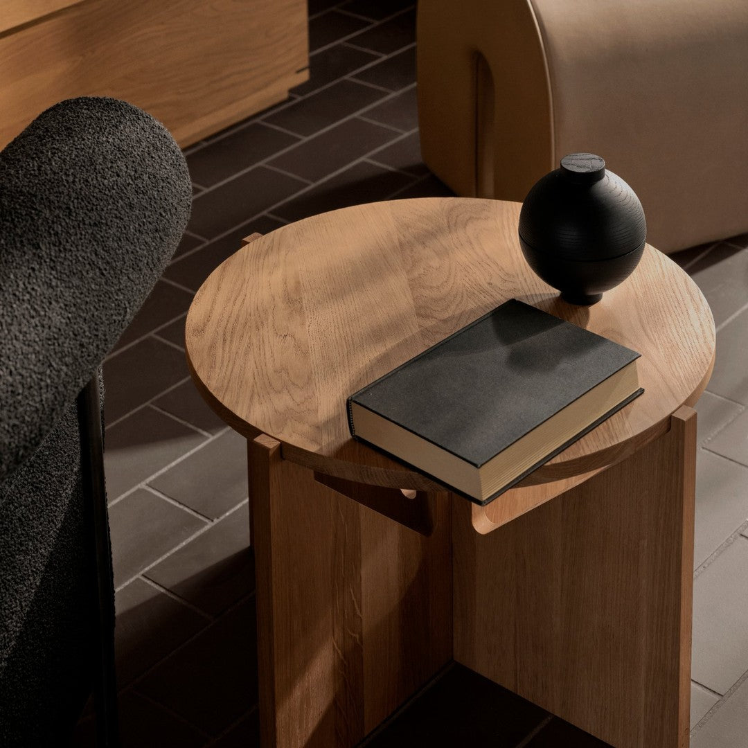 "Round wood side table with a minimalist black sphere and dark book on a textured tile floor, natural tones and sleek shapes."