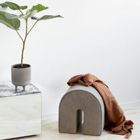 Gray arch-shaped stool with a smooth finish, complemented by a rust-colored blanket and green plant in a minimalistic setting.