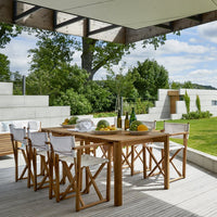 "Wooden patio dining table with white canvas chairs, surrounded by green foliage and a modern outdoor setting."