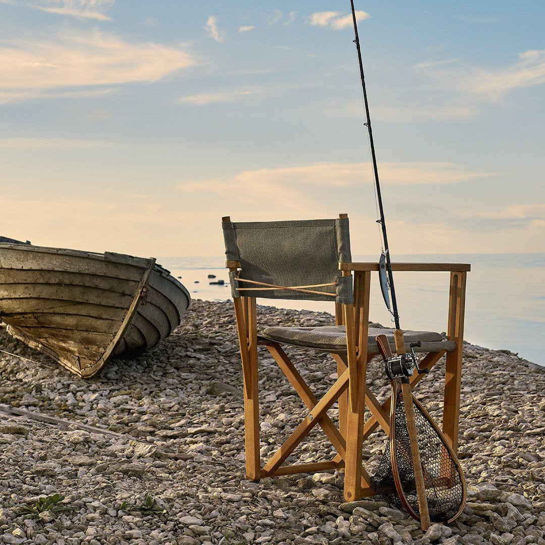 "Wooden fishing chair with a muted green canvas seat, set against a serene lakeside backdrop under a blue sky."