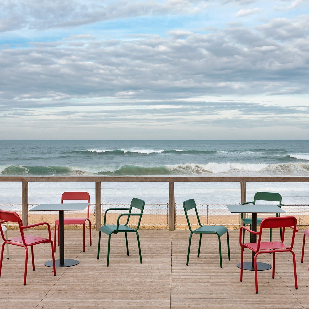 Colorful red and green chairs on a wooden deck, with waves and cloudy sky in the background. Perfect for coastal decor.
