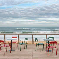 Colorful red and green chairs on a wooden deck, with waves and cloudy sky in the background. Perfect for coastal decor.