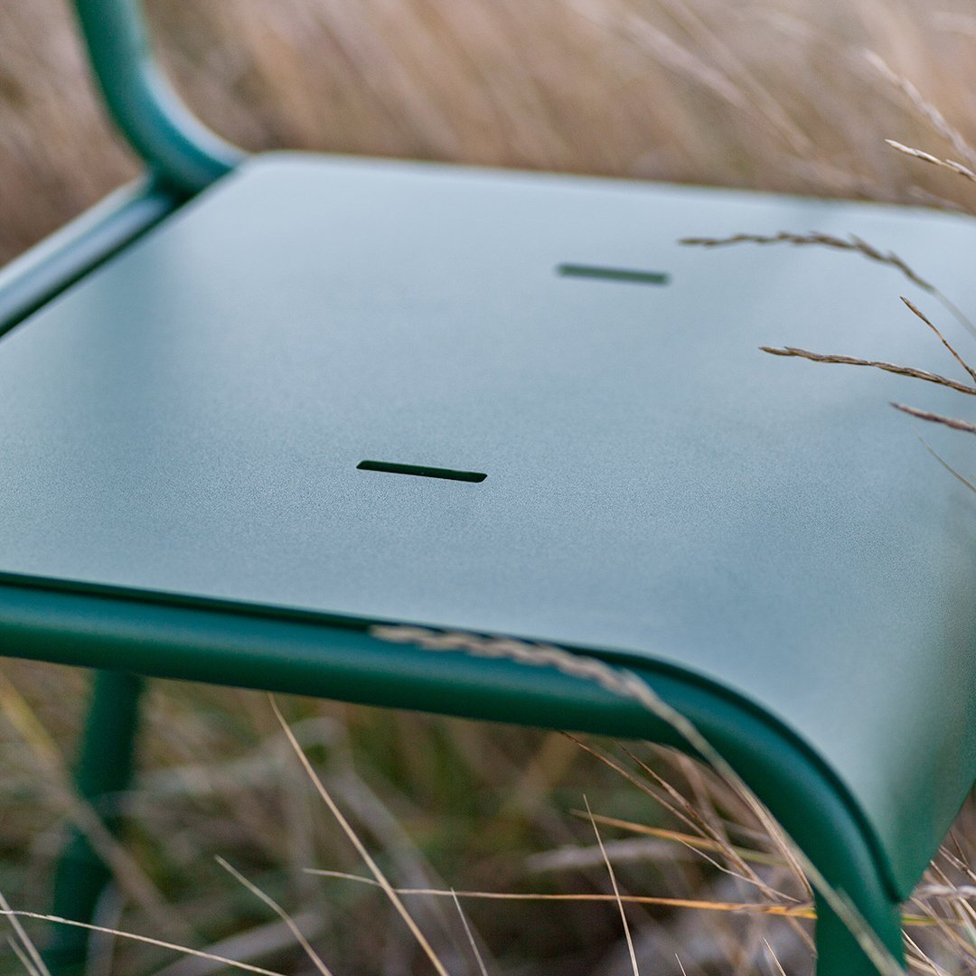 Green metal chair seat with a smooth finish and two horizontal cutouts, surrounded by soft grasses.