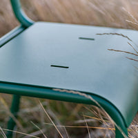 Green metal chair seat with a smooth finish and two horizontal cutouts, surrounded by soft grasses.