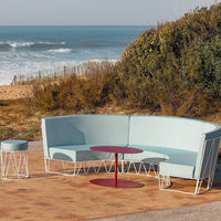 "Light blue curved sofa with white metal legs and a red circular table, set against a beach backdrop."