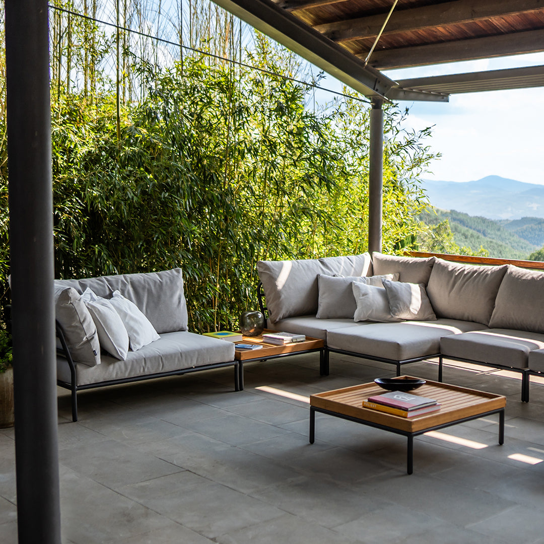 Light gray sectional sofa with white cushions, set on a gray stone patio, against lush green foliage and clear skies.