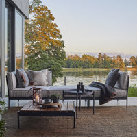 Gray outdoor sofa with patterned cushions, black coffee table, and a natural fiber rug against a serene lakeside backdrop.