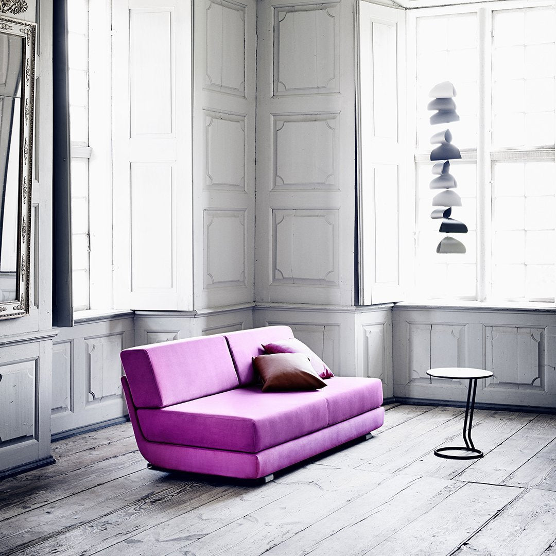 Vibrant purple couch with a simple design, accented by a dark cushion, set against a light and airy room.