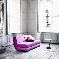 Vibrant purple couch with a simple design, accented by a dark cushion, set against a light and airy room.