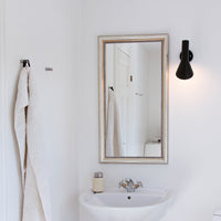 Elegant white bathroom with a black sconce, silver-framed mirror, and soft beige towels hanging. Minimalist design.