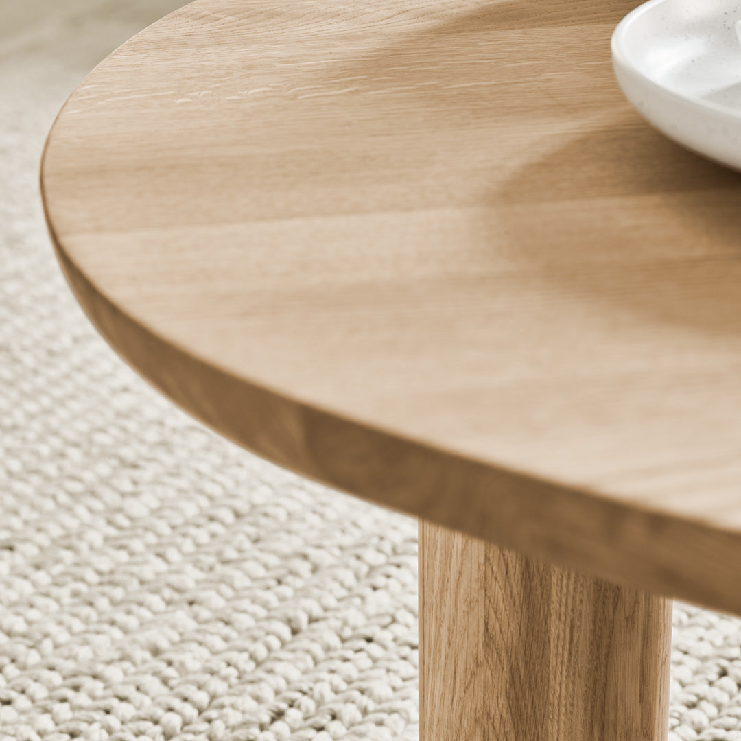 Light oak wood table with a smooth circular surface, complemented by a white dish and textured beige rug underneath.