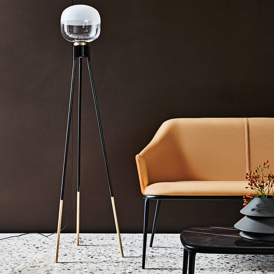 Black and gold tripod floor lamp with a spherical glass shade, paired with a warm orange modern sofa against a dark backdrop.