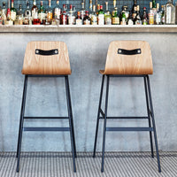 "Two modern bar stools with natural wood seats and black metal legs against a textured concrete backdrop."