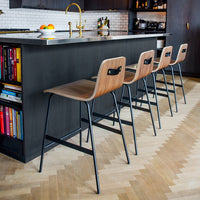 Four modern bar stools featuring warm wood seats and sleek black metal legs, set against a stylish kitchen backdrop.