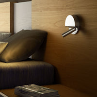 Sleek chrome wall lamp against natural wood backdrop with neutral gray pillows and a dark blue bed cover.