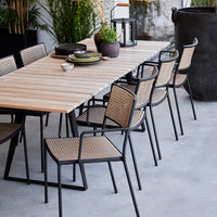 Wooden dining table with beige woven chairs and black metal frames, set in a natural outdoor setting.