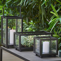 Black metal lanterns with clear glass panels, showcasing white candles and green foliage. Modern, sleek design.