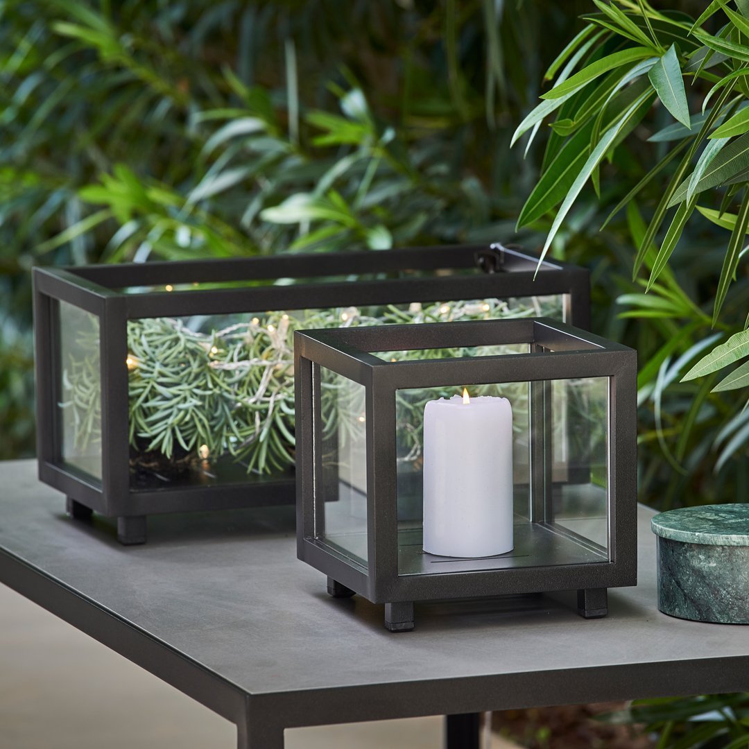 Black metal lanterns with glass panels, featuring a white candle and surrounded by lush green foliage.
