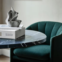 Green velvet chair with curved design alongside a black and white marbled table, featuring a gray decorative sculpture.