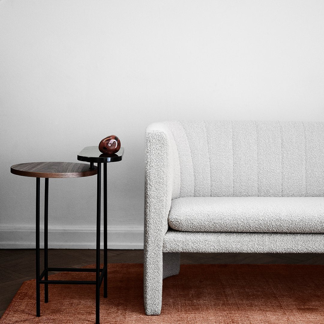 Textured white couch with subtle lines beside a round wooden side table, set on a warm orange rug. Minimalist design.