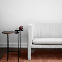Textured white couch with subtle lines beside a round wooden side table, set on a warm orange rug. Minimalist design.