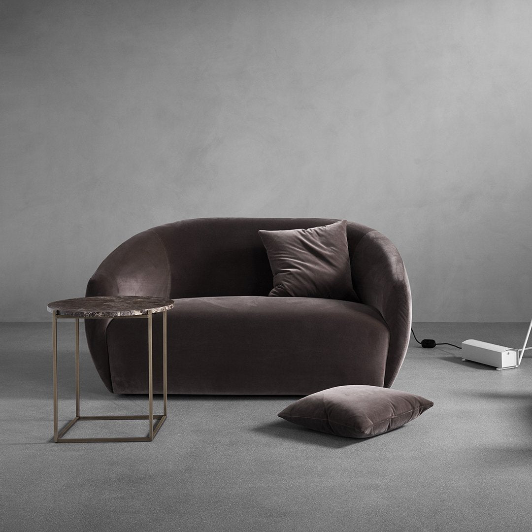 Soft gray velvet sofa with curved design, accented by a dark gray cushion and a chic gold side table. Minimalist decor elements.