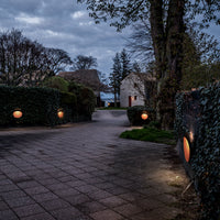 "Pathway lit by warm orange globes, surrounded by green hedges and blue-gray evening sky."