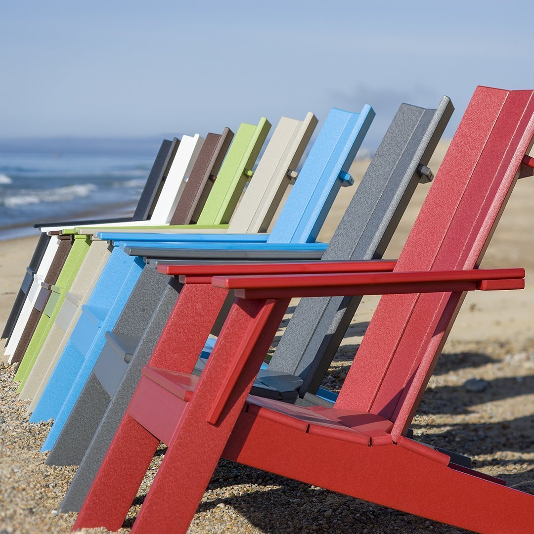 Colorful array of adirondack chairs in red, blue, green, gray, and beige, lined on a beach against a blue sky.
