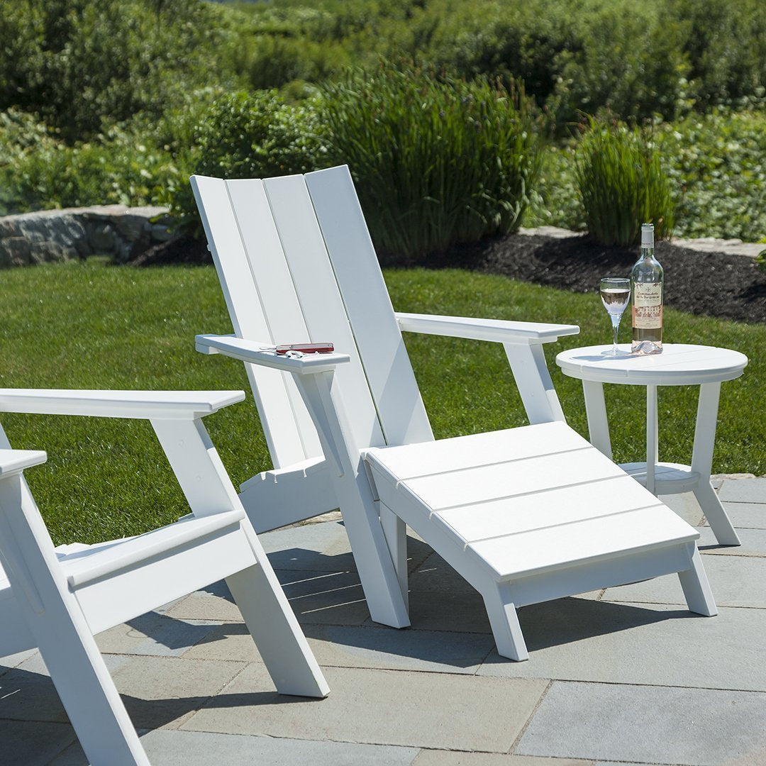 White Adirondack chairs with clean lines, paired with a round white side table on a green lawn and stone patio.