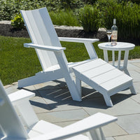 White Adirondack chair and matching side table on a stone patio, surrounded by green grass and landscaped greenery.