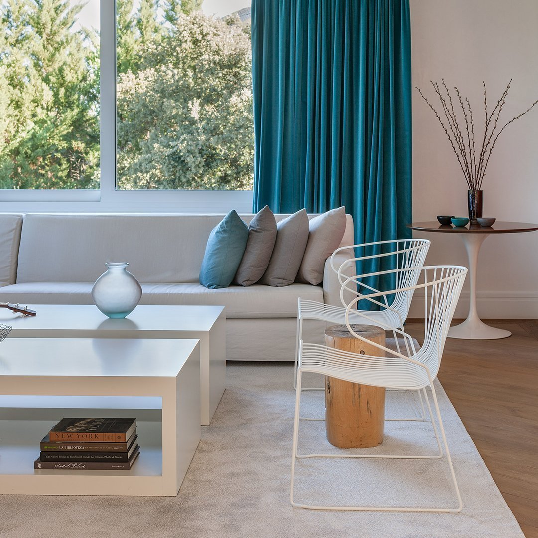 Modern living room with teal curtains, gray sofa, pastel pillows, and white chairs against a light wood floor.